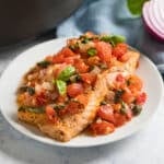 Seared salmon on a white plate with tomatoes and capers on top with the pan, a blue napkin, a piece of red onion and fresh basil behind it.