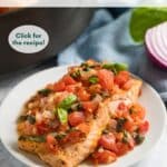 Seared salmon on a white plate with tomatoes and capers on top with the pan, a blue napkin, a piece of red onion and fresh basil behind it.