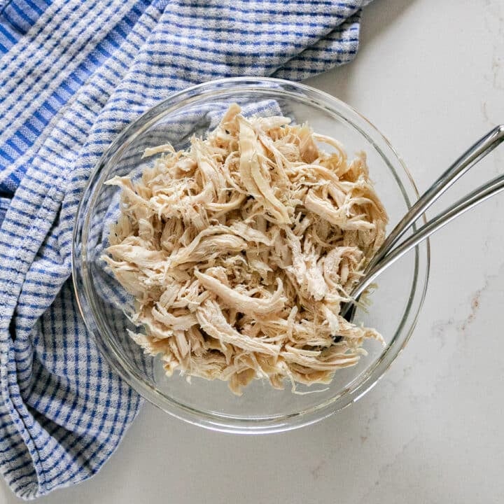 An overhead image of shredded chicken breast in a glass bowl with a blue checked towel next to it and a couple of forks in it.