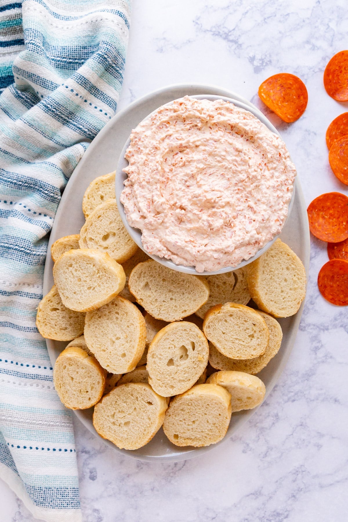 An overhead image of pepperoni dip in a bowl on a platter with baguette and some pepperoni slices and a blue striped napkin around it.