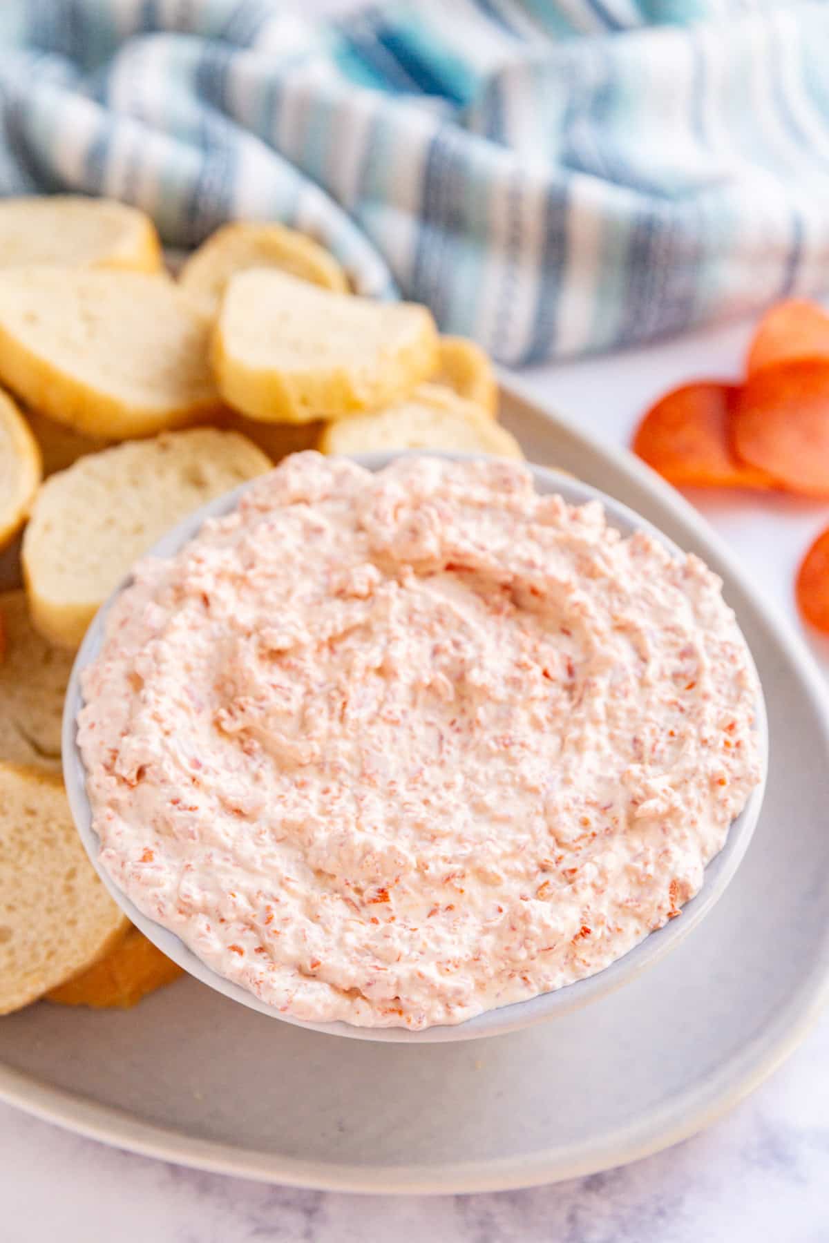 A bowl of pepperoni dip on a plate with sliced baguettes and a blue striped towel and sliced pepperoni in the background.