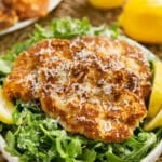 Veal scallopini on a bed of dressed arugula with lemon wedges on a white plate.