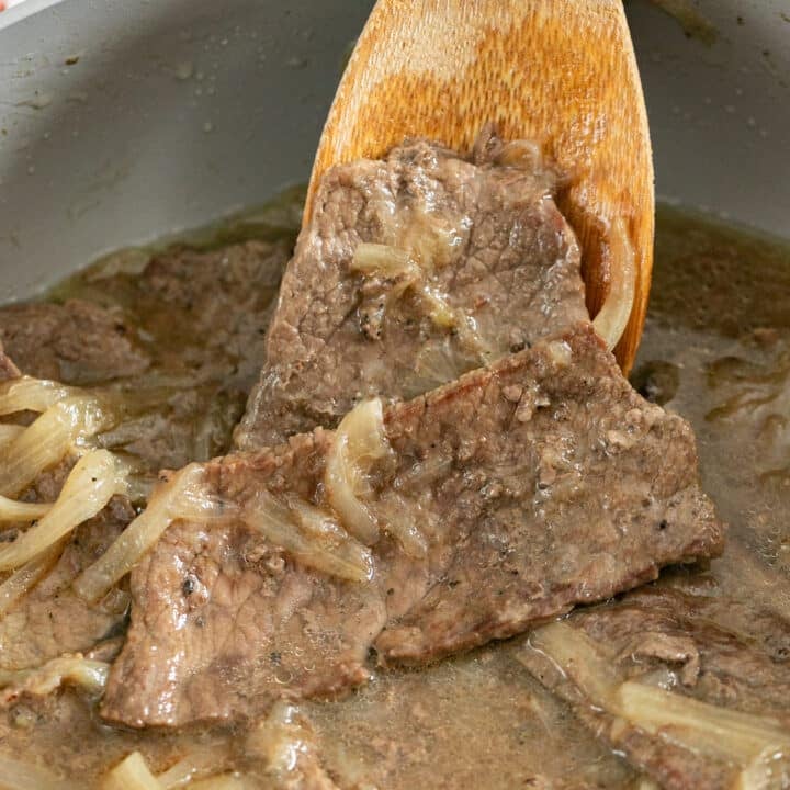 A wooden spoon resting on the side of a pan with steak and onions on it.
