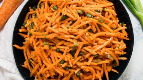 An overhead image of a dark blue bowl of carrot salad with carrots, green onion and a white napkin around it.