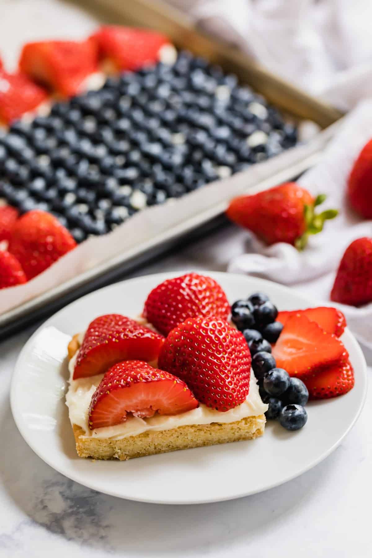 A piece of fruit pizza on a plate with the rest of it in the background.