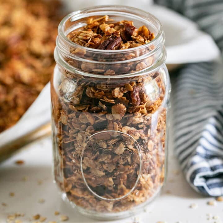 A jar with homemade granola in it with the pan of it and a napkin in the background.