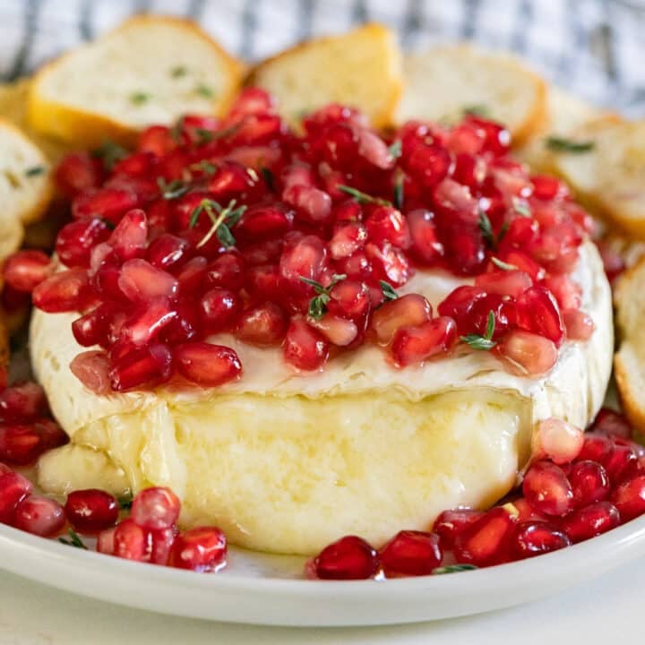 A wheel of baked brie topped with pomegranates with the front cut off and the melty cheese flowing out.