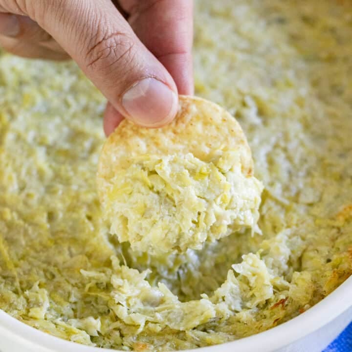 A hand pulling a chip with artichoke dip out of a pan of it.