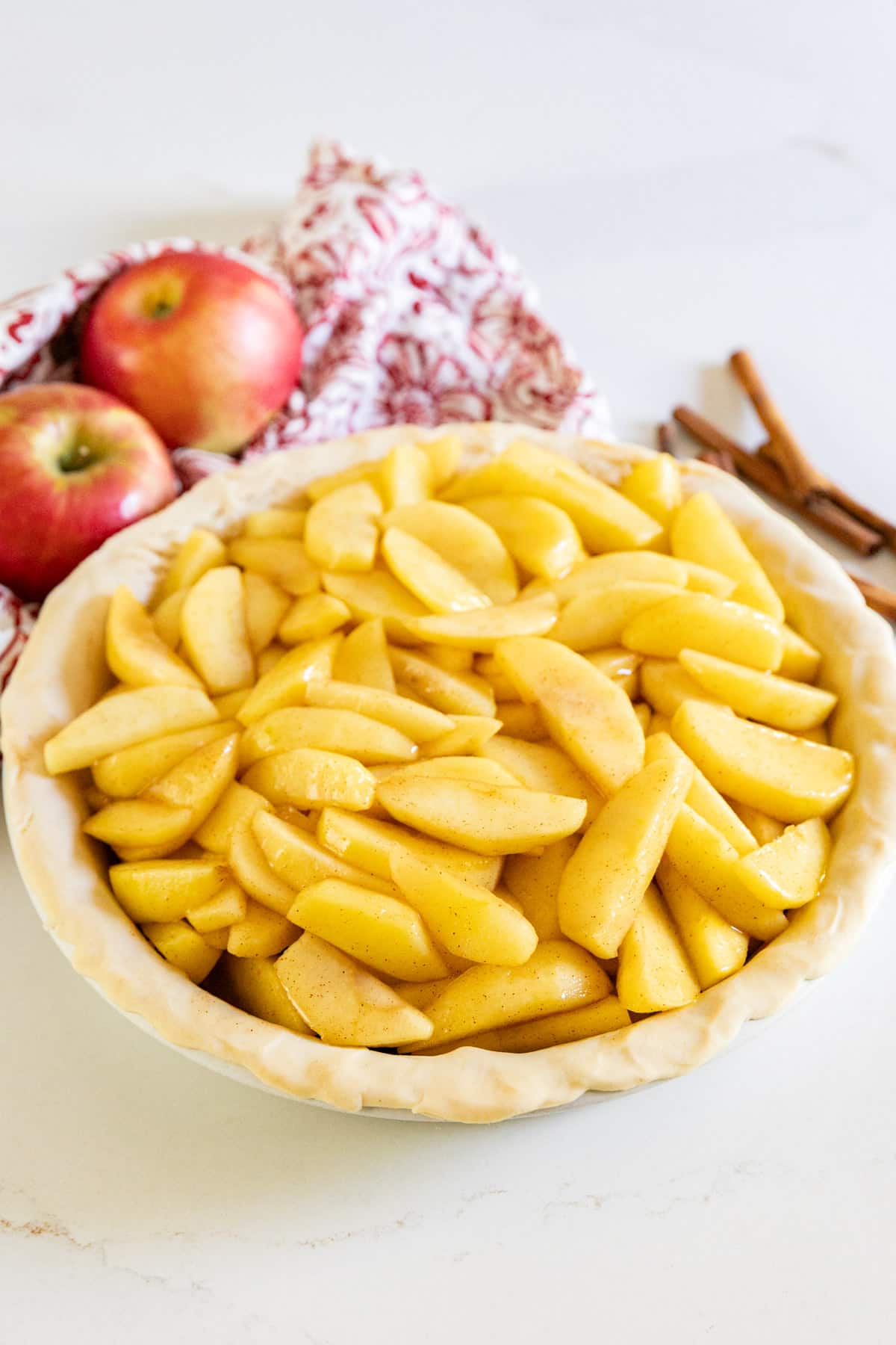 Apple pie filling in a raw pie crust with apples and cinnamon sticks behind it.