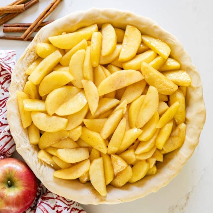 An overhead image of apple pie filling in a raw pie crust with apples and cinnamon sticks around it.