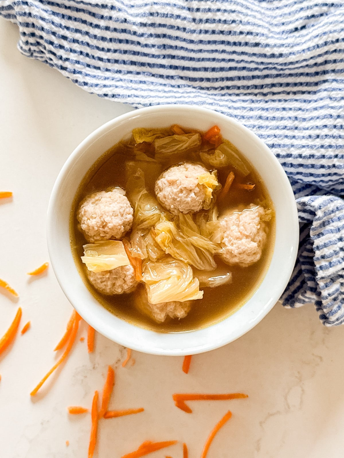 An overhead image of a bowl of Asian meatball soup with carrots and a striped napkin next to it.