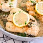 A close up of a pieces of fish in a pan of caper sauce with a lemon round on top of it.