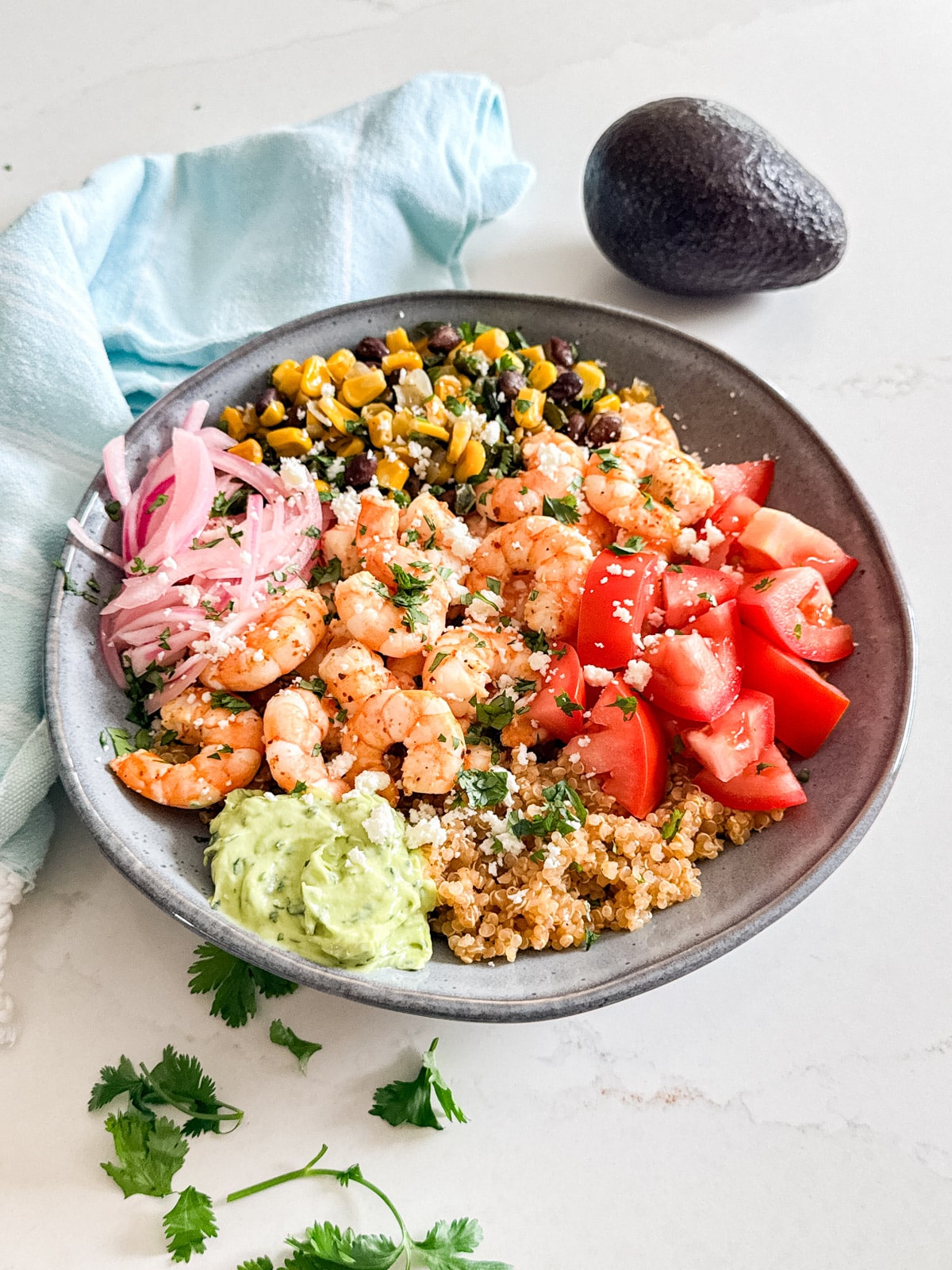 A shrimp and quinoa bowl with a towel and avocado in the background and some cilantro next to it.