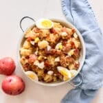 An overhead image of German potato salad on a bowl with potatoes and a blue towel next to it.