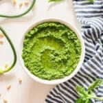 An overhead image of a bowl of garlic scape pesto with scapes and basil around it and a striped towel.